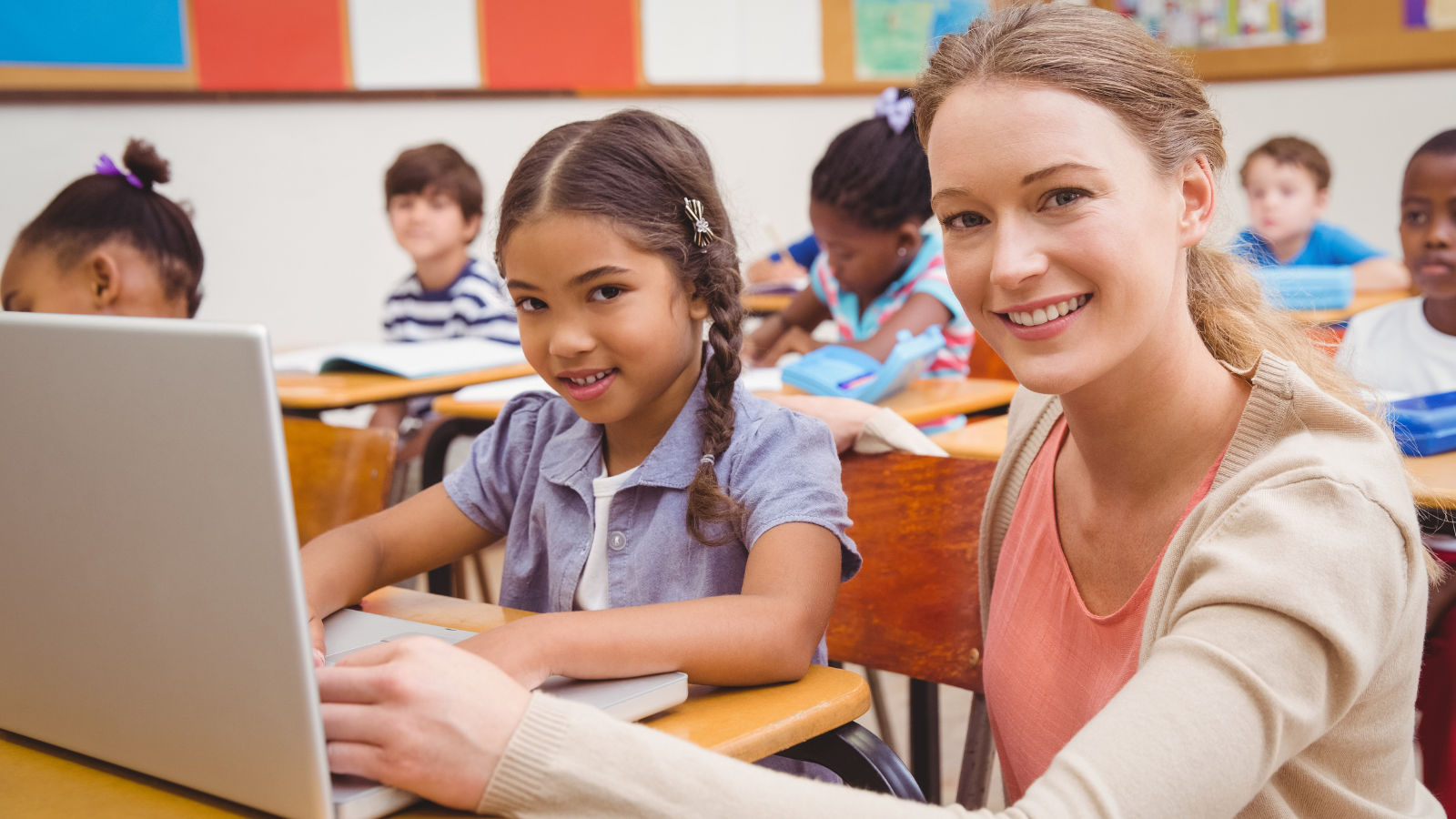 Female elementary student sitting at a desk with computer and her femail teacher sitting besider her.